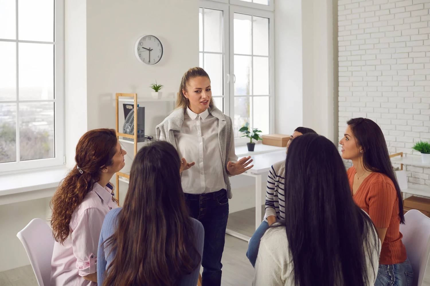 A woman stands and gestures while addressing a group of attentive listeners in a bright, modern room with large windows and plants A woman stands and gestures while addressing a group of attentive listeners in a bright, modern room with large windows and plants