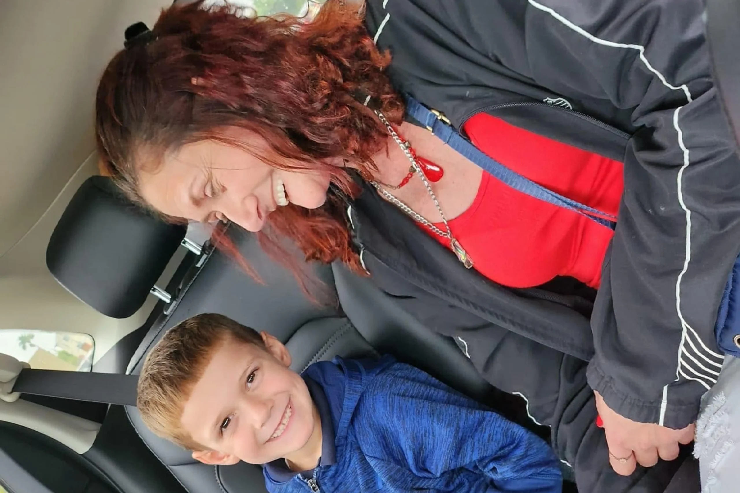 A person with curly hair wears a black jacket over a red shirt, seated in a car beside a child in a blue hoodie, both engaged with a device A person with curly hair wears a black jacket over a red shirt, seated in a car beside a child in a blue hoodie, both engaged with a device