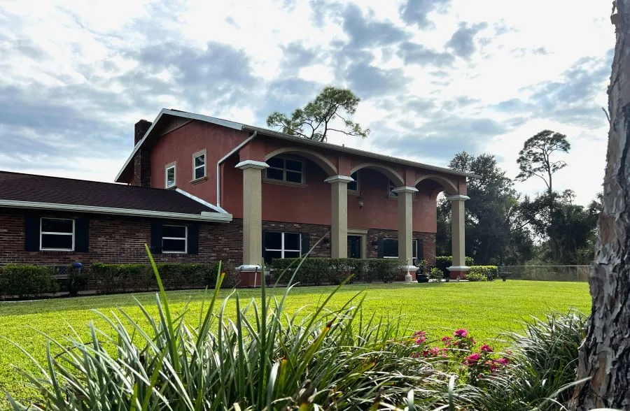 A spacious two-story house with red walls and white columns, surrounded by lush green grass and colorful flowers under a cloudy sky A spacious two-story house with red walls and white columns, surrounded by lush green grass and colorful flowers under a cloudy sky